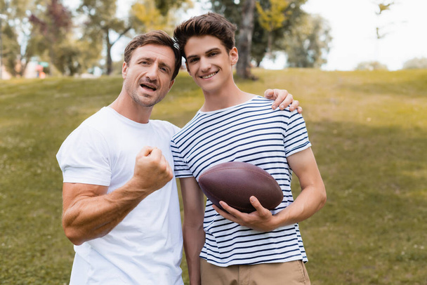 Aufgeregter Vater zeigt geballte Faust und steht mit Rugbyball neben Teenager-Sohn  - Foto, Bild