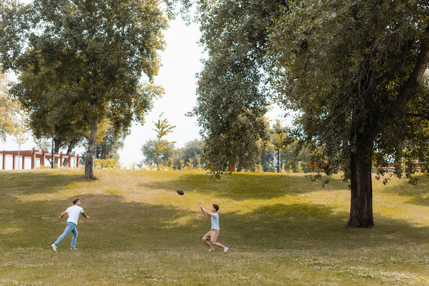 vader en tiener zoon spelen amerikaans voetbal in groen park  - Foto, afbeelding