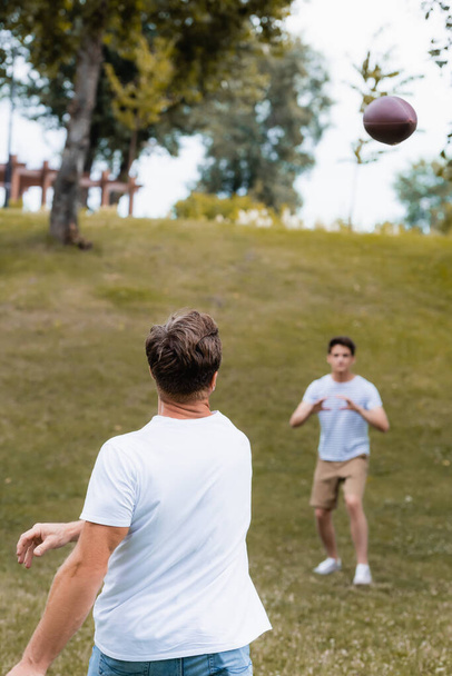 enfoque selectivo de padre lanzando pelota de rugby cerca de hijo adolescente en parque verde  - Foto, Imagen