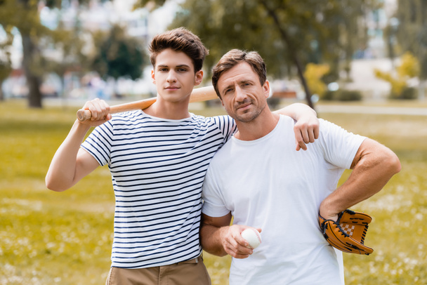 adolescente menino com morcego softball de pé perto do pai em luva de couro segurando bola  - Foto, Imagem