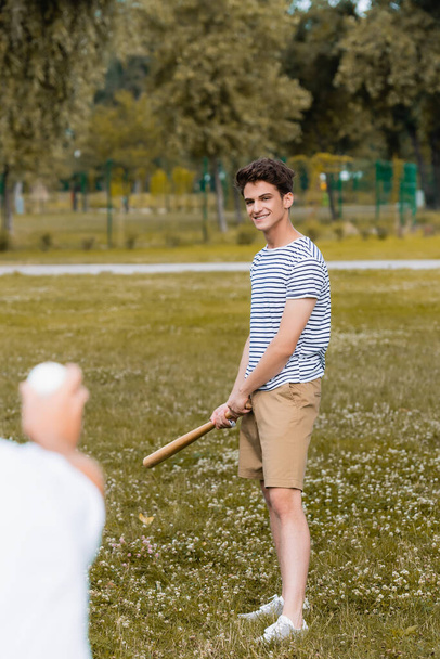 enfoque selectivo de alegre hijo adolescente sosteniendo el bate de softbol cerca del padre en el parque - Foto, imagen