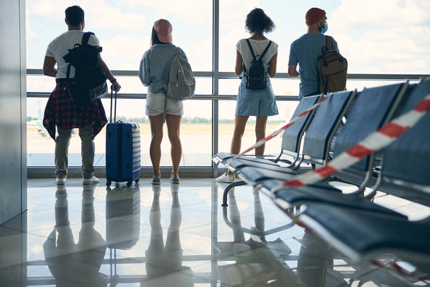 Vier mensen in casual kleren die uit het raam kijken terwijl ze op een vlucht wachten - Foto, afbeelding