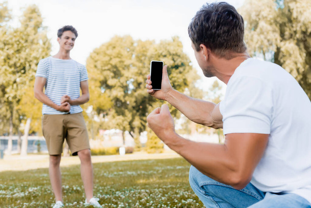 スマートフォンを持っていて公園で10代の息子の写真を撮るという選択的な焦点は  - 写真・画像