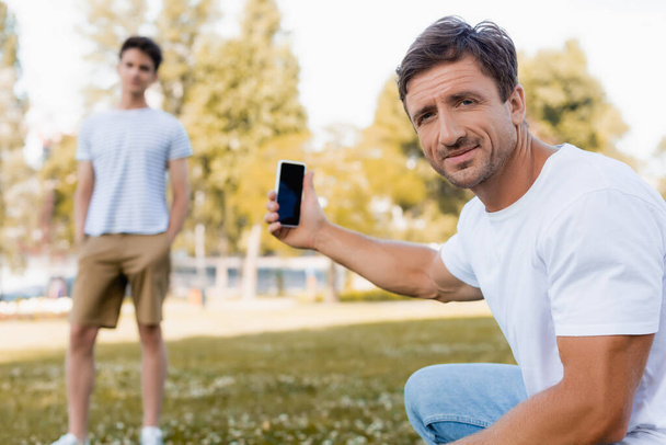 selectieve focus van vader houden smartphone met leeg scherm in de buurt van tiener zoon in het park  - Foto, afbeelding