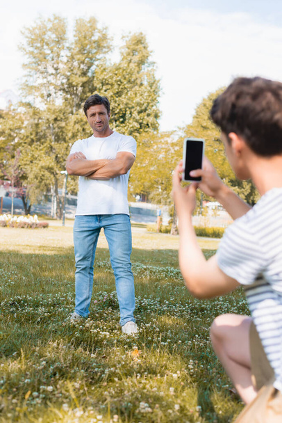 10代の男の子が公園で写真を撮りスマートフォンを持っている間に交差腕で立っている男の選択的な焦点  - 写真・画像