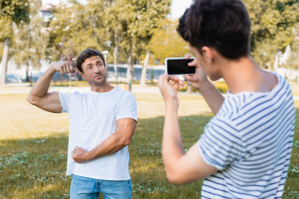 selective focus of strong father posing while teenager boy taking photo and holding smartphone in park  - Фото, изображение
