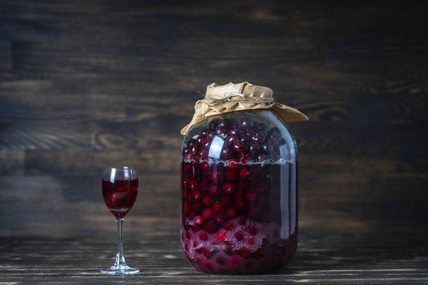 Kotitekoinen tinktuura punaisesta kirsikasta. Berry alkoholijuomien käsite. Kotitekoinen punaviini valmistettu kypsistä kirsikoista suuressa lasipurkissa ja viinilasi puupohjalla, Ukraina - Valokuva, kuva