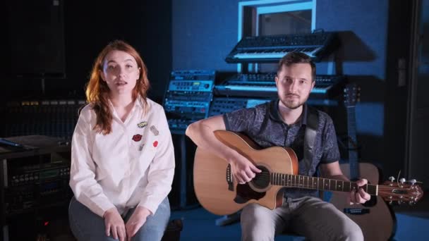 Guitarrista guapo con barba acompaña a una chica pelirroja en una guitarra en un estudio de música. Cámara de mano. Mediano tiro largo - Metraje, vídeo