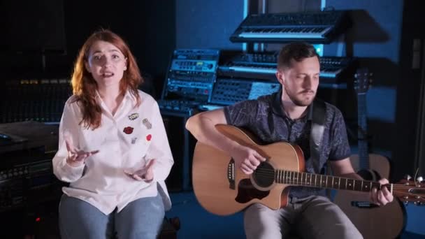 Der gutaussehende Gitarrist mit Bart begleitet ein rothaariges Mädchen auf der Gitarre in einem Musikstudio. Handkamera. Mittlerer Weitschuss - Filmmaterial, Video