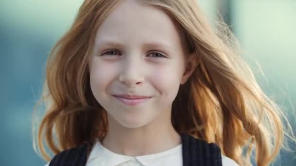 Portret klein gelukkig schattig blond school meisje met blauwe ogen ziet er vriendelijk op camera glimlachende tanden staan op straat buiten, genieten van de kindertijd, wind speelt met lang goud haar, onschuld concept - Video