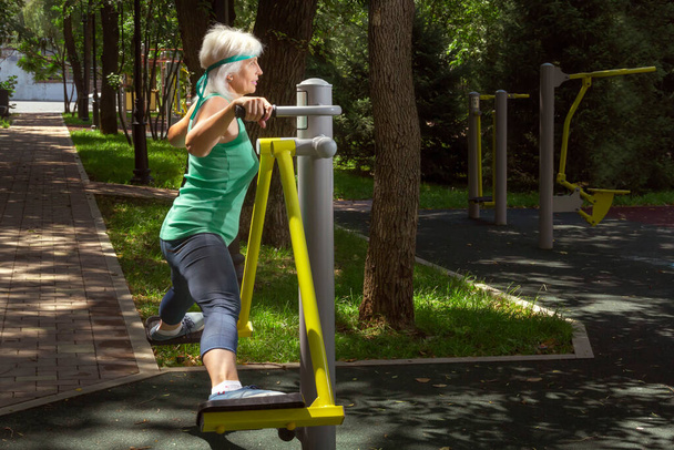Mulher idosa magra com cabelo grisalho conduz aulas de fitness individuais em um simulador em um parque da cidade. Espaço de cópia - Foto, Imagem