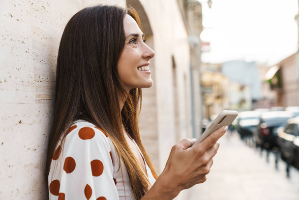Image of happy brunette woman smiling and using mobile phone while walking on city street - Foto, Imagen