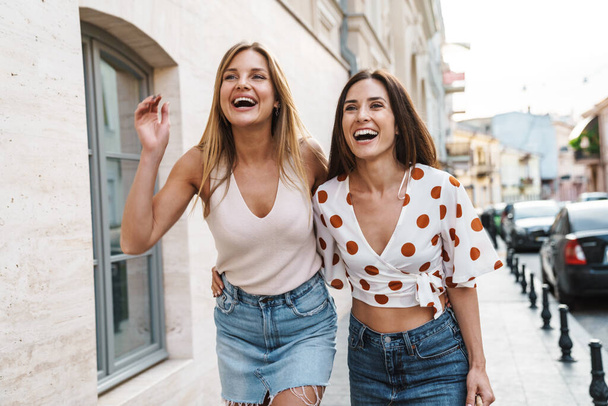 Image of cheerful adult two women hugging and laughing while walking on city street - Photo, image