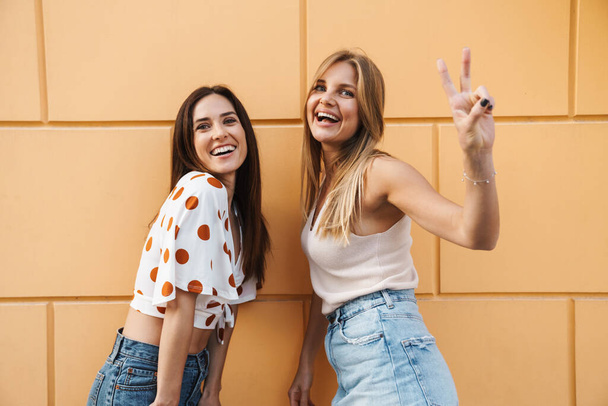 Image of excited adult two women laughing and gesturing peace sign while posing on city street - Fotó, kép