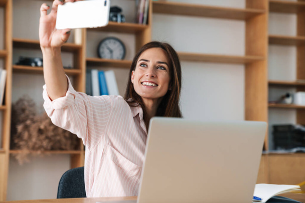 Image of happy businesswoman smiling and taking selfie on cellphone while working with laptop in office - Photo, Image