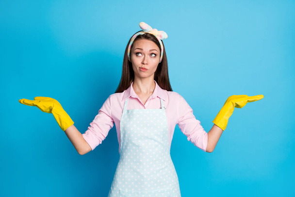 Portrait of unsure minded girl hold hand present adverts promo think thoughts hesitate wear pink shirt yellow latex gloves headband isolated blue color background - Photo, image