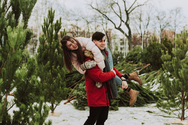 若いカップルは街のクリスマスツリー市場を背景に冬を楽しんで、周りをばかにしています。彼氏の後ろに若い女が笑って笑っている. - 写真・画像