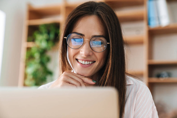 Image closeup of happy adult businesswoman in eyeglasses smiling while working with laptop at office - Φωτογραφία, εικόνα