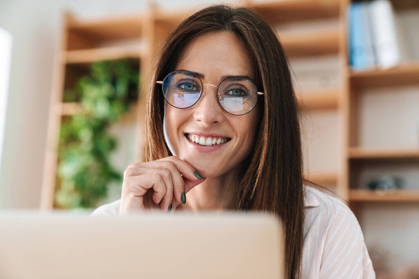 Image closeup of happy adult businesswoman in eyeglasses smiling while working with laptop at office - Фото, изображение