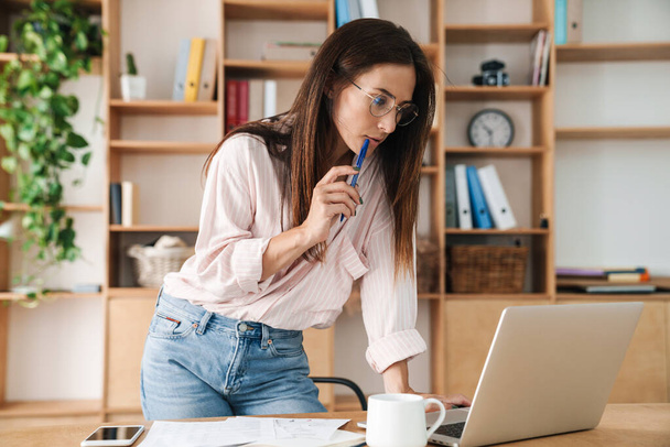 Image of concentrated adult businesswoman in eyeglasses working with laptop at office - Zdjęcie, obraz