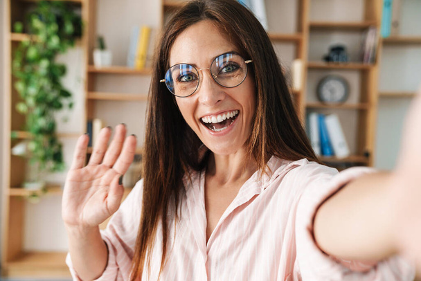 Image of excited adult businesswoman waving hand while taking selfie photo in office - Fotografie, Obrázek