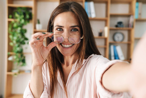 Зображення щасливої дорослої бізнес-леді посміхається і тримає окуляри, роблячи селфі фото в офісі
 - Фото, зображення