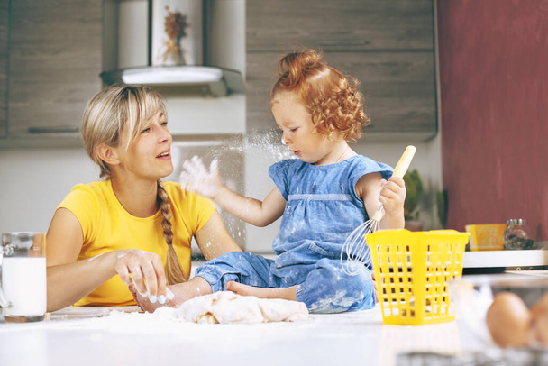 Een jonge mooie vrouw en haar dochtertje kneden koekjesdeeg in de keuken. Het meisje houdt een garde in haar handen en speelt met bloem, zittend. Familie vrijetijdsbesteding, recreatie samen - Foto, afbeelding