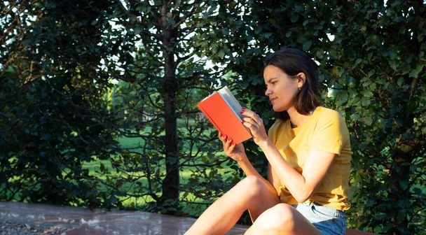 Junges erwachsenes kaukasisches Mädchen im gelben T-Shirt sitzt morgens im Park und liest Buch - Foto, Bild