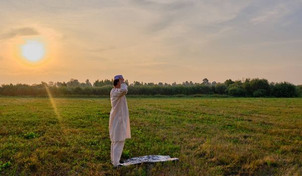 A Muslim senior man wearing an embroidered skullcap and white traditional clothes prays at sunset in a field Banner - Fotoğraf, Görsel