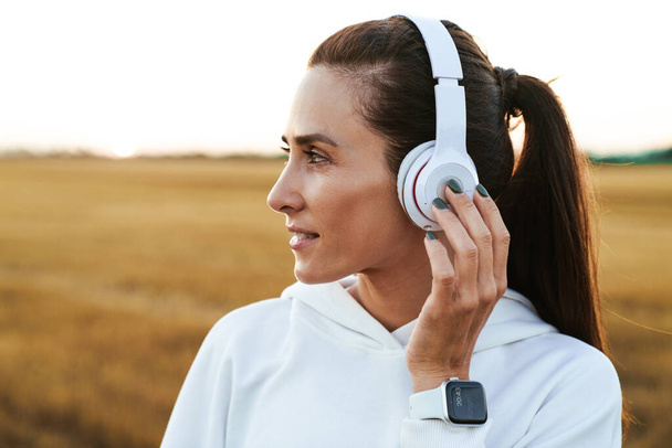 Image of pleased sportswoman in smartwatch using wireless headphones while working out at golden field - Zdjęcie, obraz