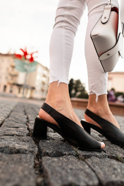 ファッショナブルな黒革の夏の靴で白いデニムパンツの女性の足のクローズアップは、通りにスタイリッシュなバッグ. - 写真・画像