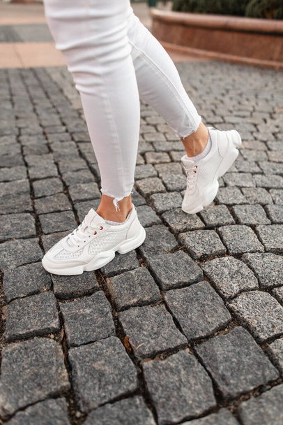 Fashion female legs in white sneakers and denim pants on a stone road in the city. Close-up. - Photo, image