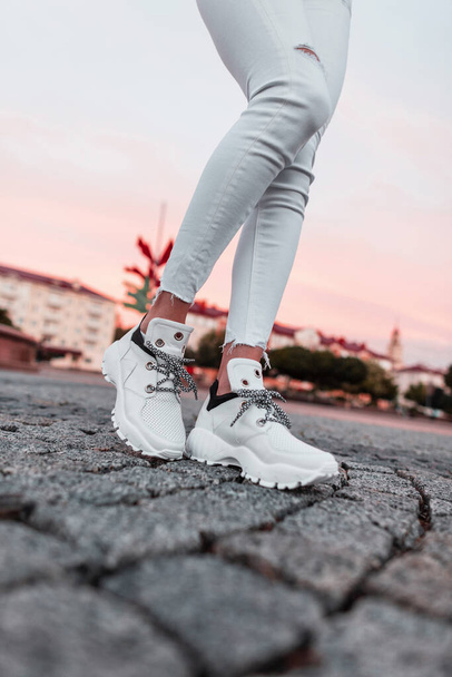 Fashionable casual white sneakers on female legs on a stone road at pink sunset. Close-up. - Foto, Imagen