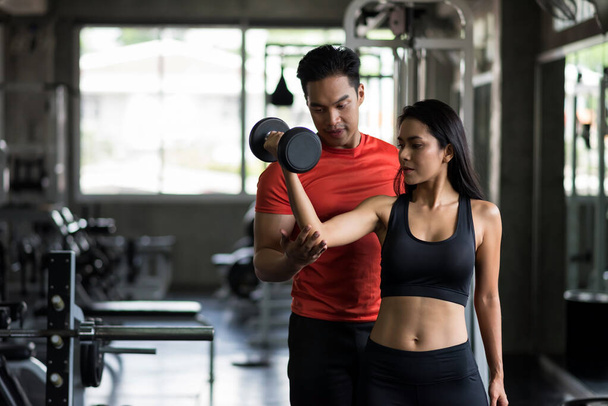 Personal fitness trainer or boyfriend coaching dumbbell exercise by adjust elbow and arm to sexy woman in sport gym. Happy couple working out for bodybuilding and healthy lifestyle. - Photo, Image