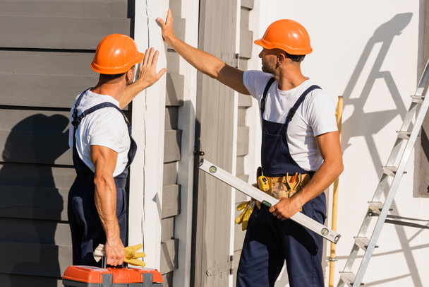 Constructores en uniforme y hardhats tocando fachada de edificio al aire libre  - Foto, Imagen