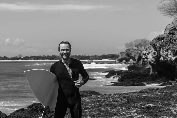 Black and white portrait of handsome man surfer in black wetsuit, holding white surf board  and blue ocean, cliff, rocks on background - Fotografie, Obrázek