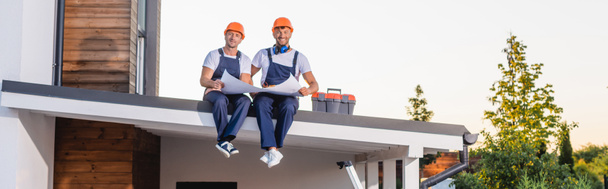 Panoramaaufnahme von Bauarbeitern in Uniform, die einen Bauplan in der Nähe des Werkzeugkastens auf dem Dach des Gebäudes halten  - Foto, Bild
