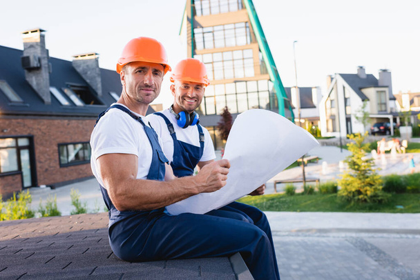 Arbeiter in Overalls halten Bauplan auf Hausdach in der Stadtstraße  - Foto, Bild