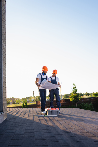 Builders with blueprint standing near toolbox on roof of building  - Photo, Image