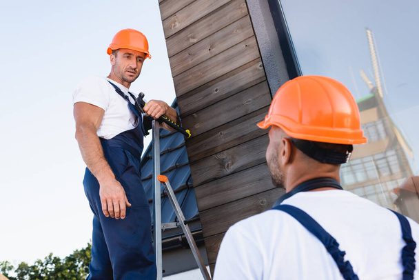 Вибіркове фокусування будівельника, який тримає молоток, стоячи на драбині біля колеги і фасаду будівництва  - Фото, зображення