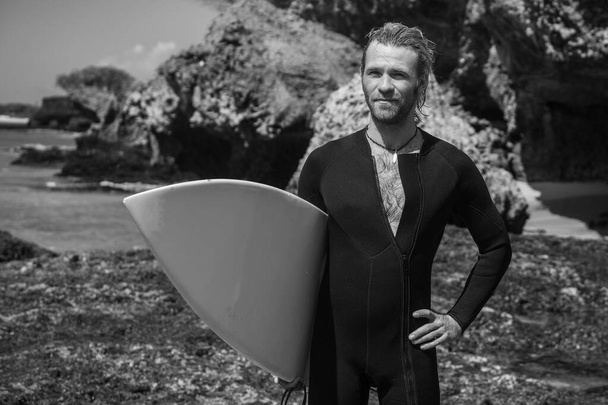 Black and white portrait of handsome man surfer in black wetsuit, holding white surf board  and blue ocean, cliff, rocks on background - Foto, Imagen