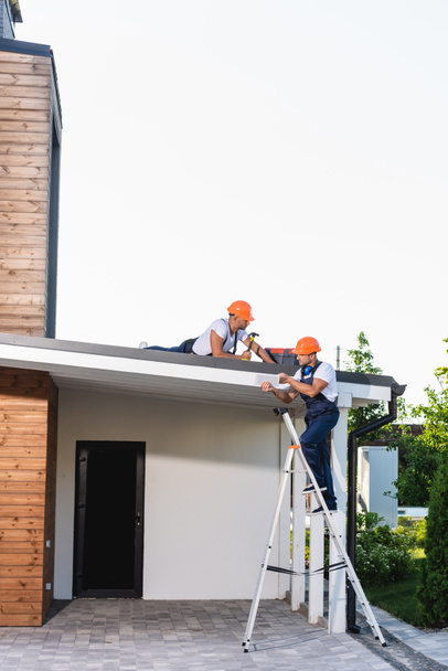 Construtor na escada segurando tablet digital perto colega com martelo no telhado da casa  - Foto, Imagem