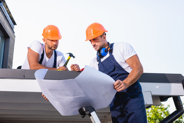 Handyman holding blueprint while standing on ladder near colleague with hammer on roof of building  - Photo, Image