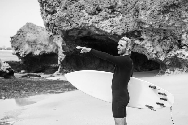 Black and white portrait of handsome man surfer in black wetsuit, holding white surf board  and blue ocean, cliff, rocks on background - Foto, Imagem