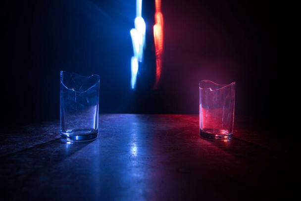 Pij, stłuczone szkło. Stłuczone szklanki na drewnianym stole w ciemnym, stonowanym tle z mgłą. Skupienie selektywne - Zdjęcie, obraz