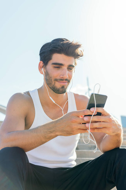 vertical photo of a handsome sportsman with earphones listening to music and consulting his phone, concept of technology and healthy lifestyle, copy space for text - Fotografie, Obrázek