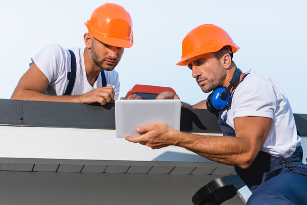 建物の屋根の上で作業しながら、デジタルタブレットを使用して作業中の手仕事の男性の選択的な焦点  - 写真・画像