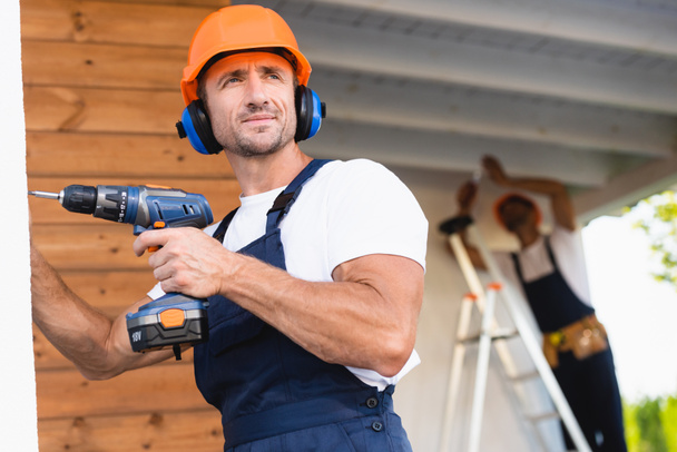 Selective focus of handyman in workwear and ear defenders holding electric screwdriver near facade of building  - Photo, Image