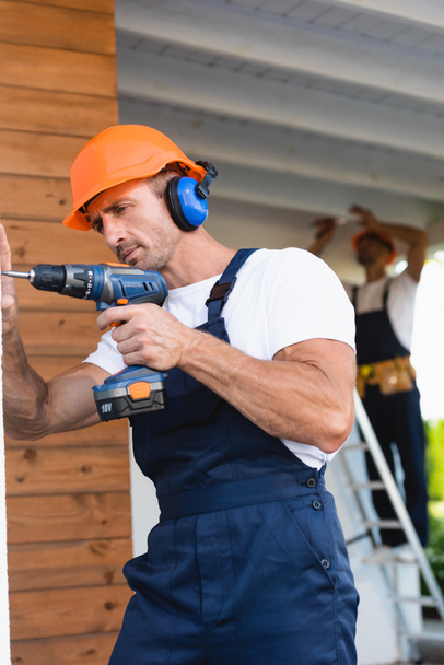 Selective focus of manual worker in helmet and ear defenders holding electric screwdriver near facade of house  - Photo, Image