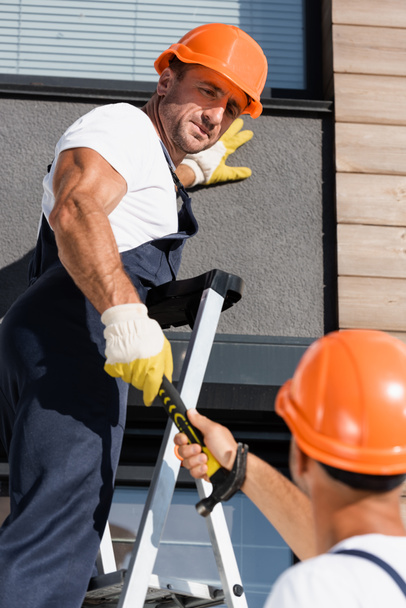 Selektiver Fokus des Bauherrn gibt Hammer an Kollegen auf Leiter nahe Hausfassade ab  - Foto, Bild